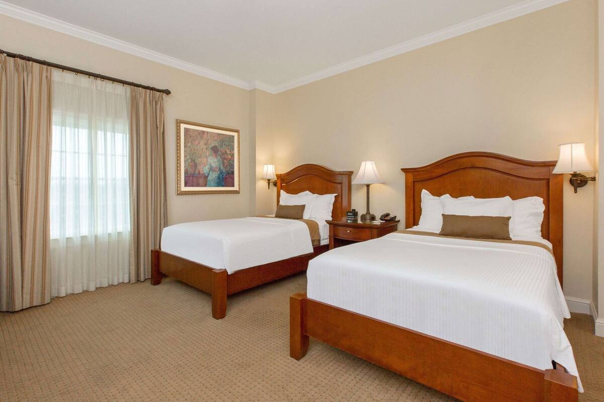 A hotel room with two single beds, a large window with sheer curtains, a painting on the wall, and a nightstand with a lamp.