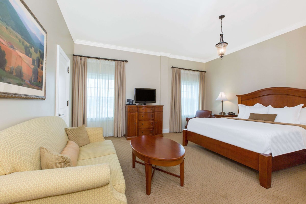 A hotel room with a bed, a sofa, a TV, a dresser, and a painting on the wall.
