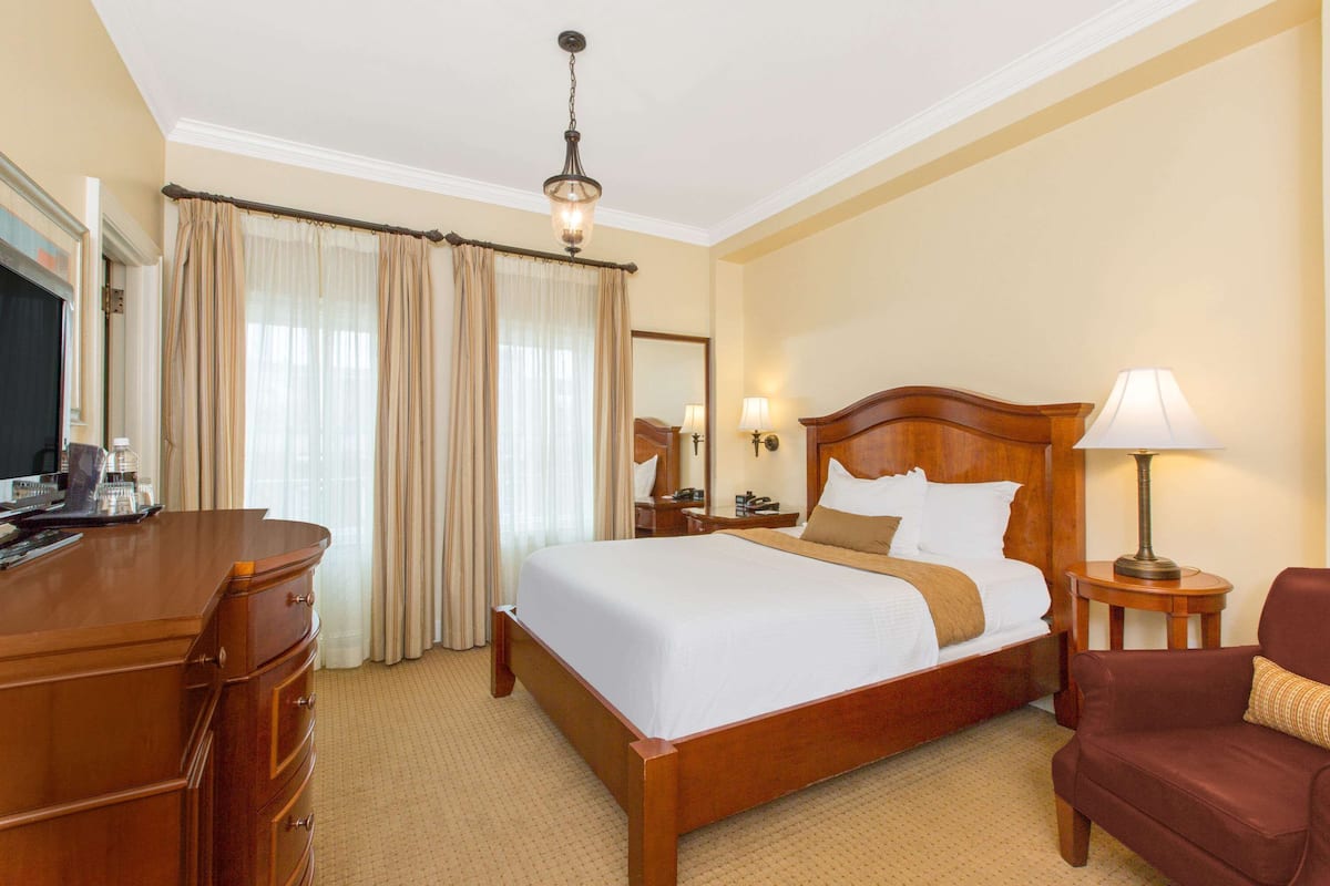 A hotel room with a large bed, wooden headboard, a desk with a lamp, a chair, and a dresser.