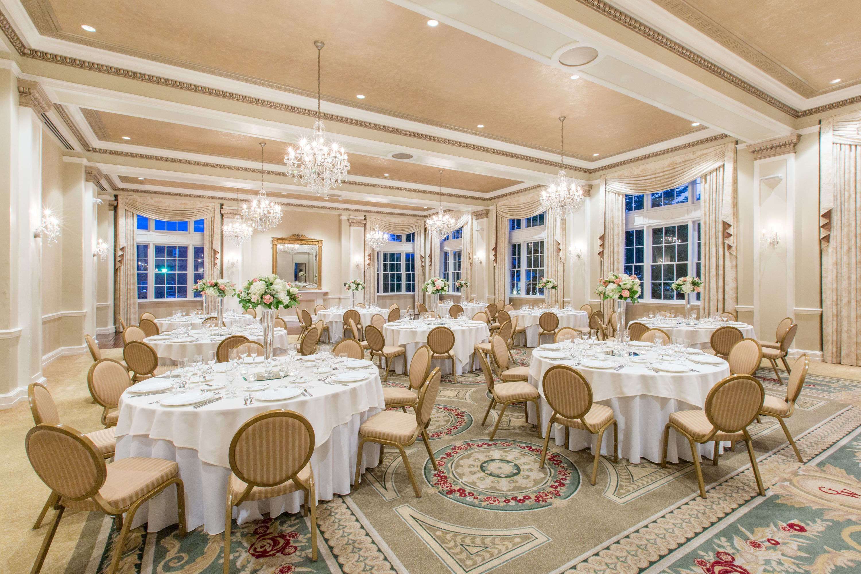 A banquet hall with round tables, chairs, chandeliers, and floral centerpieces.