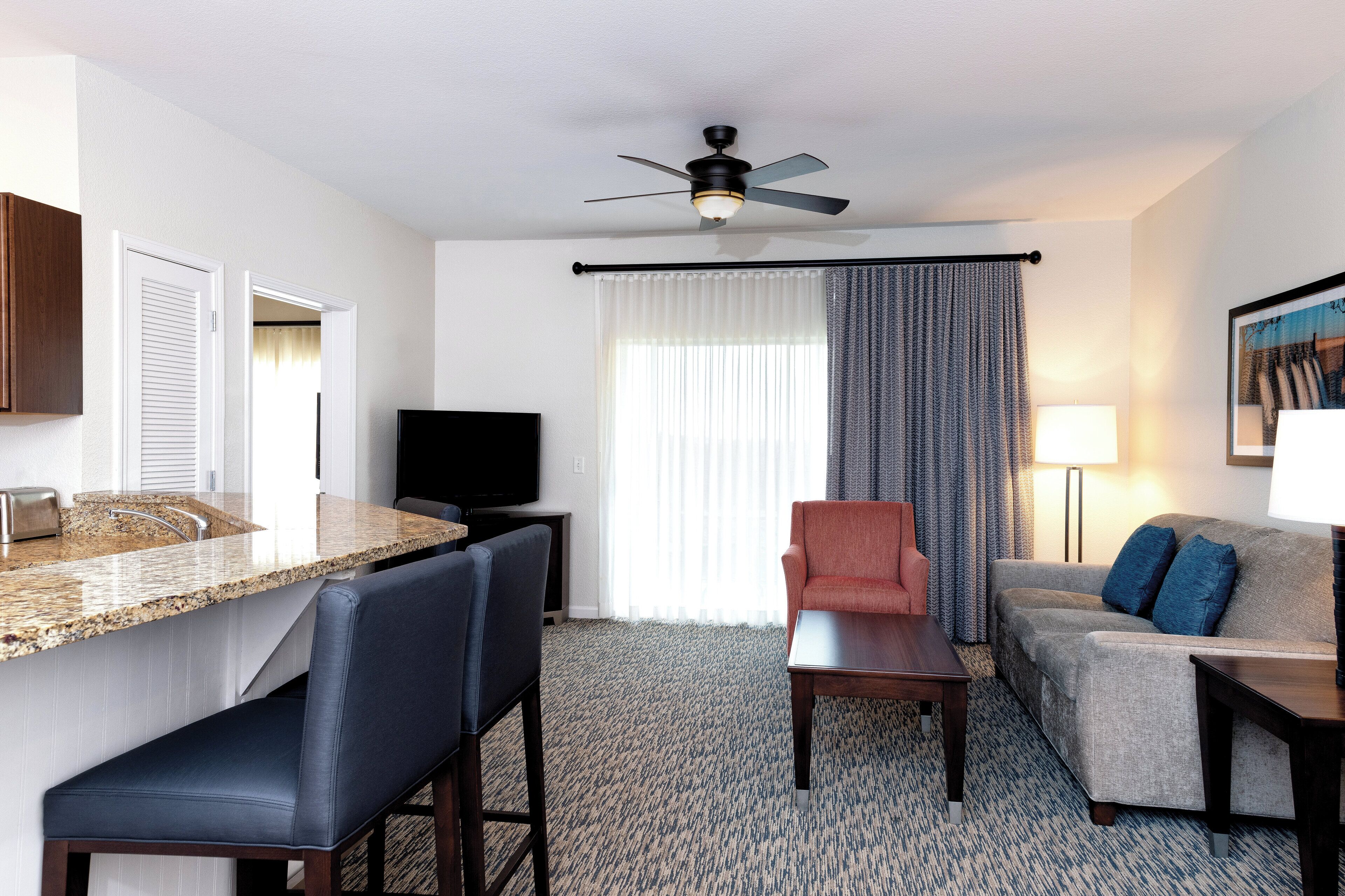 Quarto de hotel com cozinha compacta, sofá, poltrona, televisão e ventilador de teto.