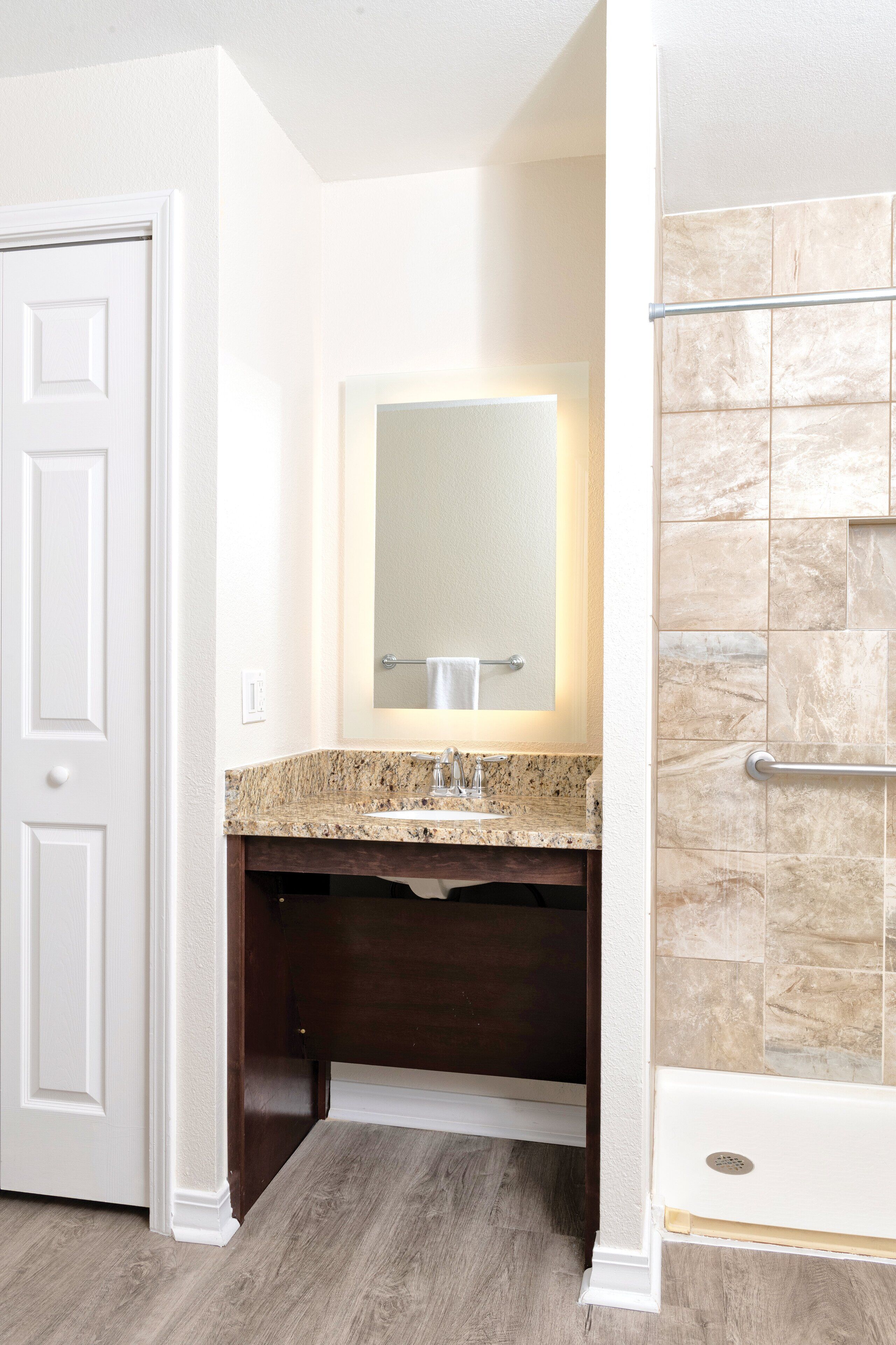 Une salle de bain avec une porte blanche, un lavabo doté d’un plan de travail en granit, un miroir et une douche aux carreaux beiges.