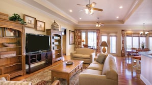 A spacious living room with a flat-screen TV, comfortable seating, wooden furniture, and a ceiling fan.
