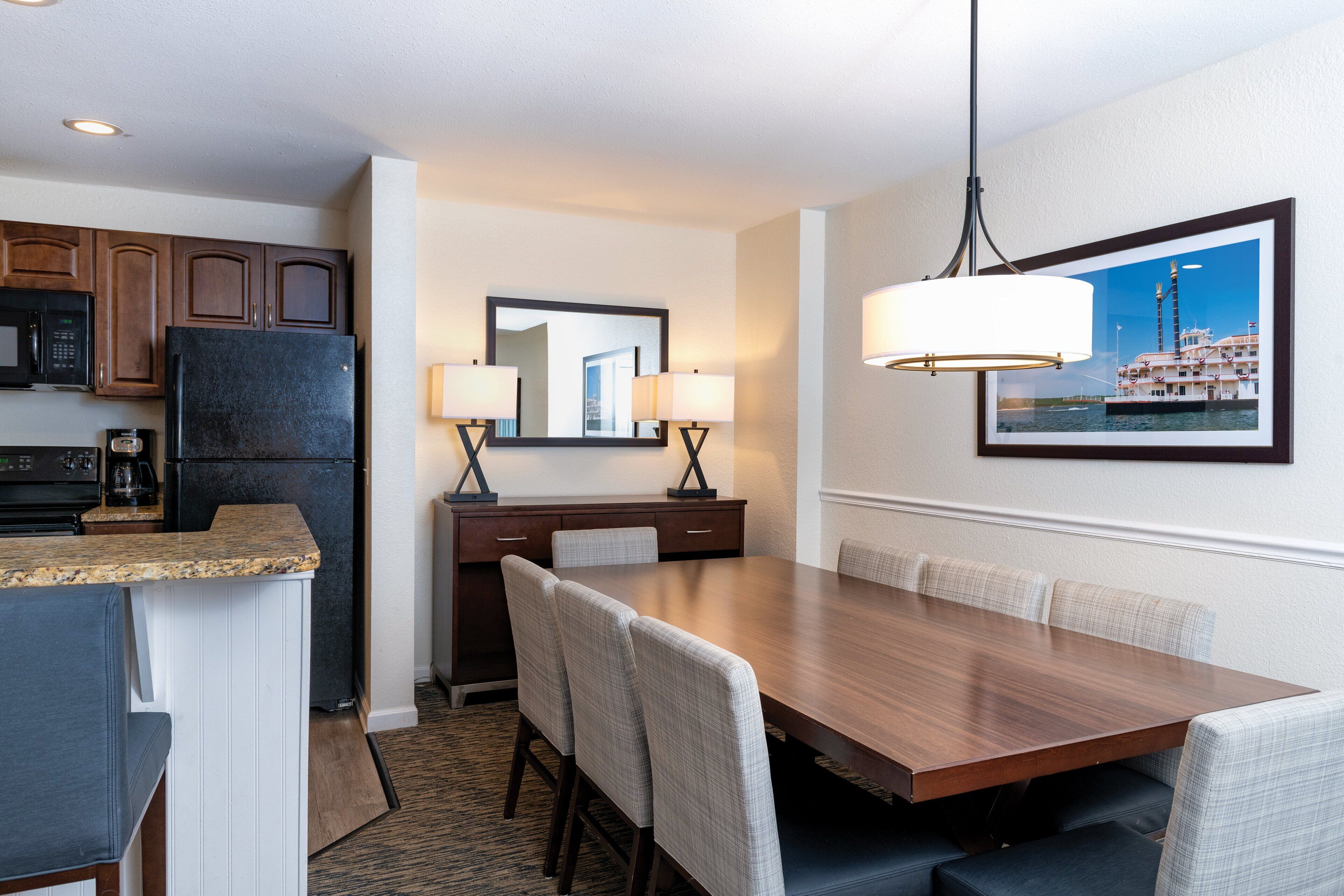 A hotel room with a dining area featuring a wooden table, cushioned chairs, a microwave, a refrigerator, and a framed picture on the wall.