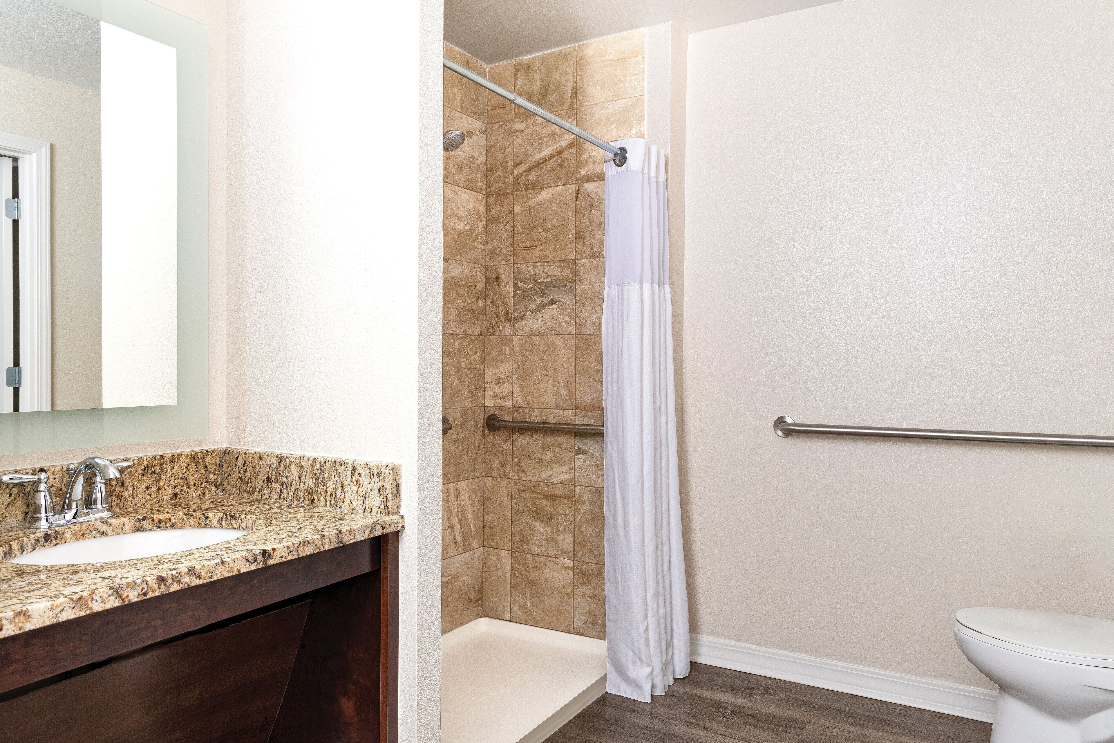 Une salle de bain avec un lavabo sur un plan de travail en granit, des toilettes blanches, une douche avec un rideau blanc et une barre d’appui.