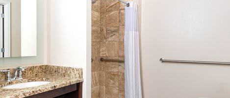Une salle de bain avec un lavabo sur un plan de travail en granit, des toilettes blanches, une douche avec un rideau blanc et une barre d’appui.