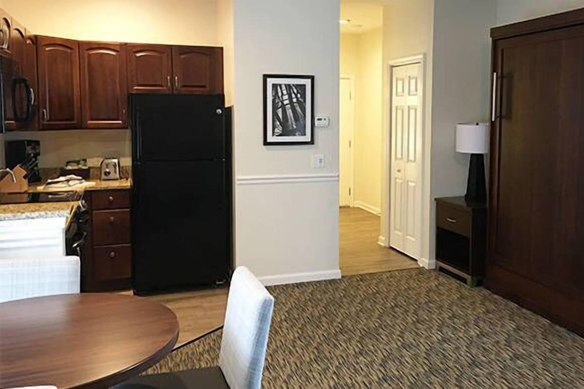Una cocina con armarios de madera, un refrigerador negro, una mesa de comedor con sillas y piso alfombrado.