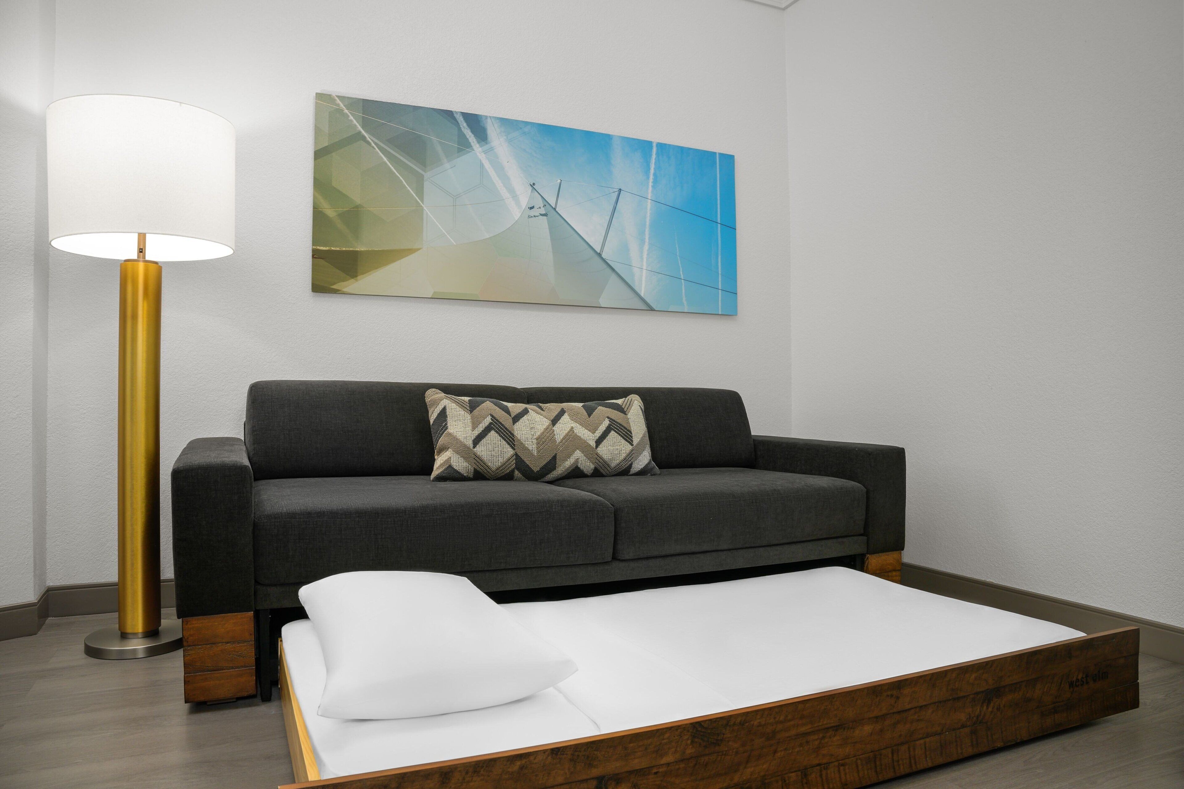 A modern room with a grey sofa, a white mattress, a floor lamp, and a geometric wall art piece.