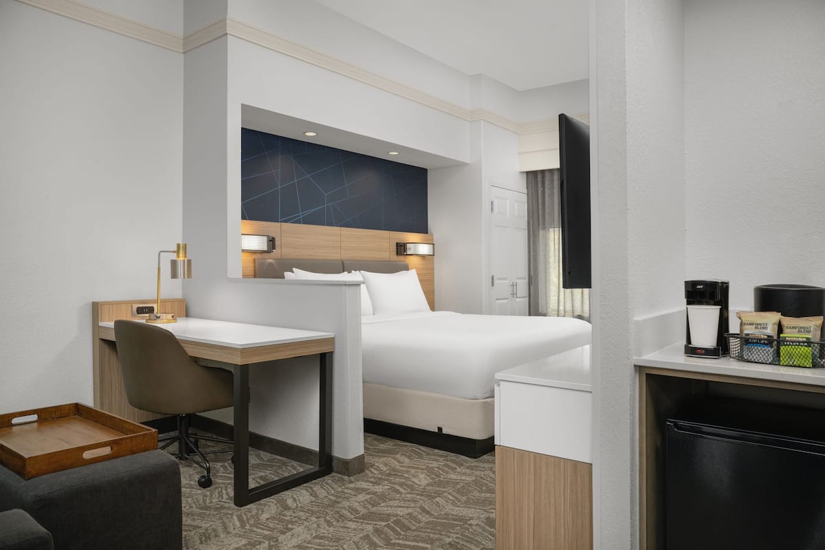 A hotel room with a bed, a desk, a chair, a coffee machine, and a tray table.