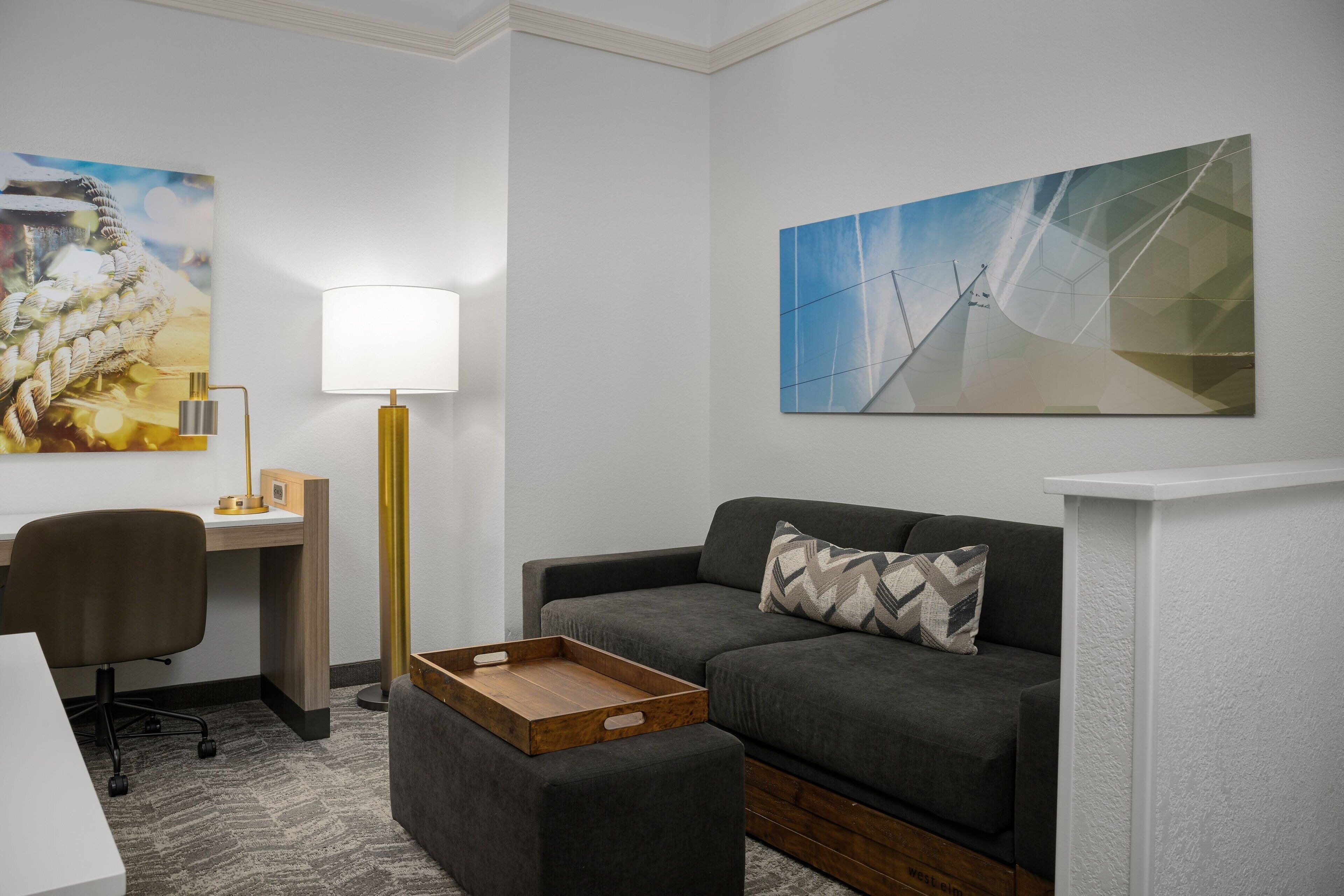 Une chambre d’hôtel moderne avec un canapé gris, une table d’appoint en bois, un bureau avec une lampe et deux œuvres d’art accrochées aux murs.