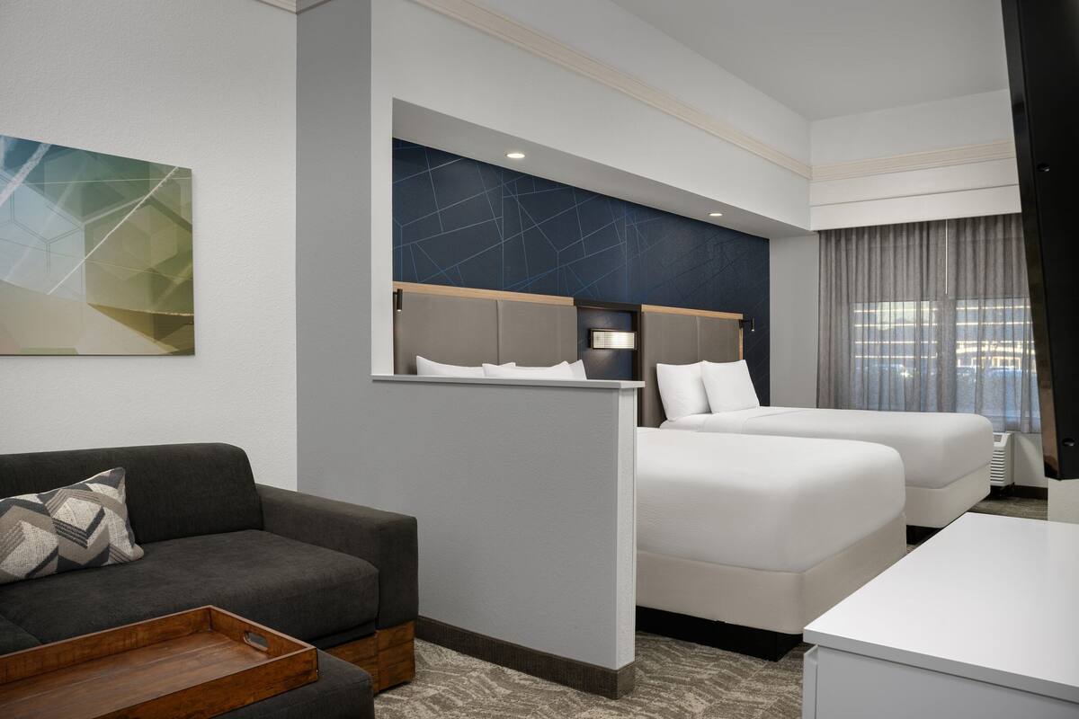 A hotel room with a gray sofa, a bed with white bedding, a desk, and a large window with curtains.