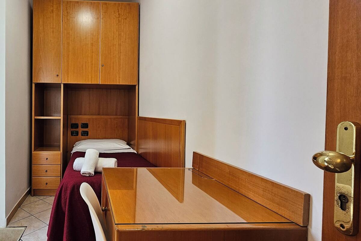 A small, compact room with a bed, a wooden wardrobe, and a small desk.