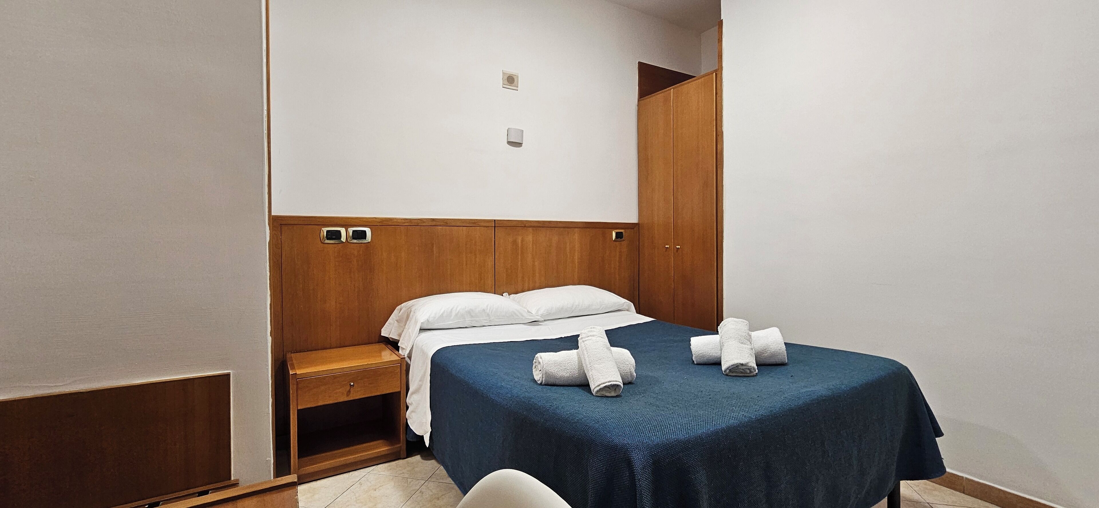 A hotel room with a bed, a wooden wardrobe, a nightstand, and a small table.