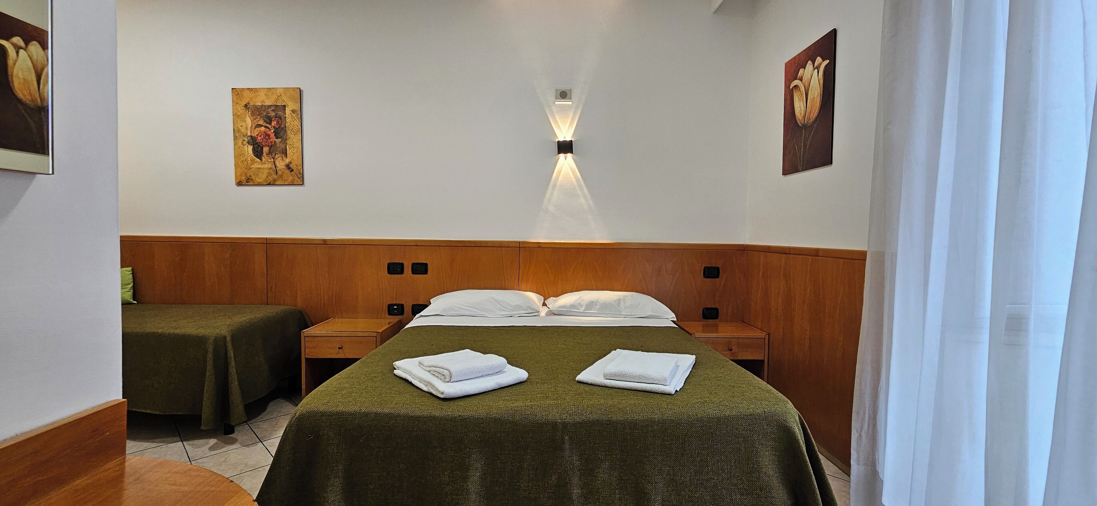 Habitación de hotel con una cama, dos mesitas de noche, una lámpara de pared y dos cuadros.