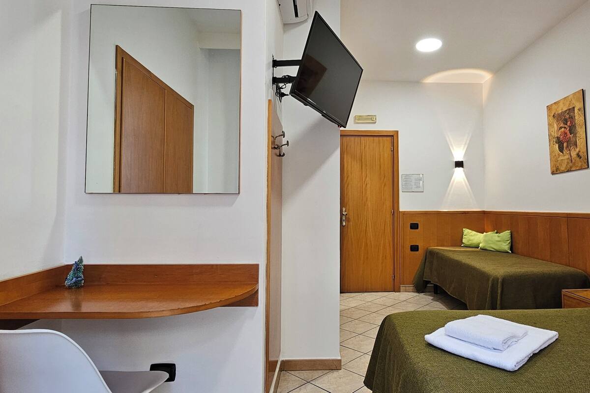 A hotel room with a wooden desk, a television mounted on the wall, two beds with green bedspreads, and a small Christmas tree on the desk.