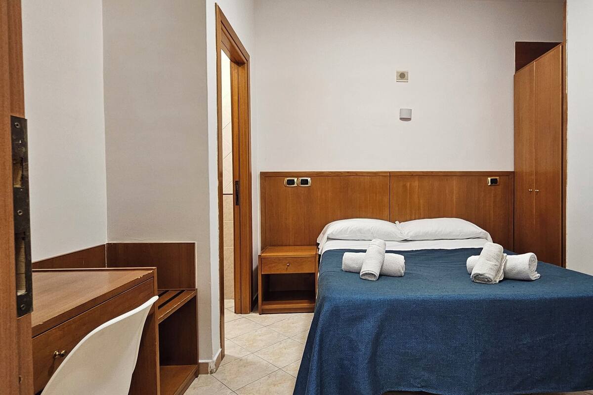 A small, neatly arranged hotel room with a bed, a desk, and a wardrobe.