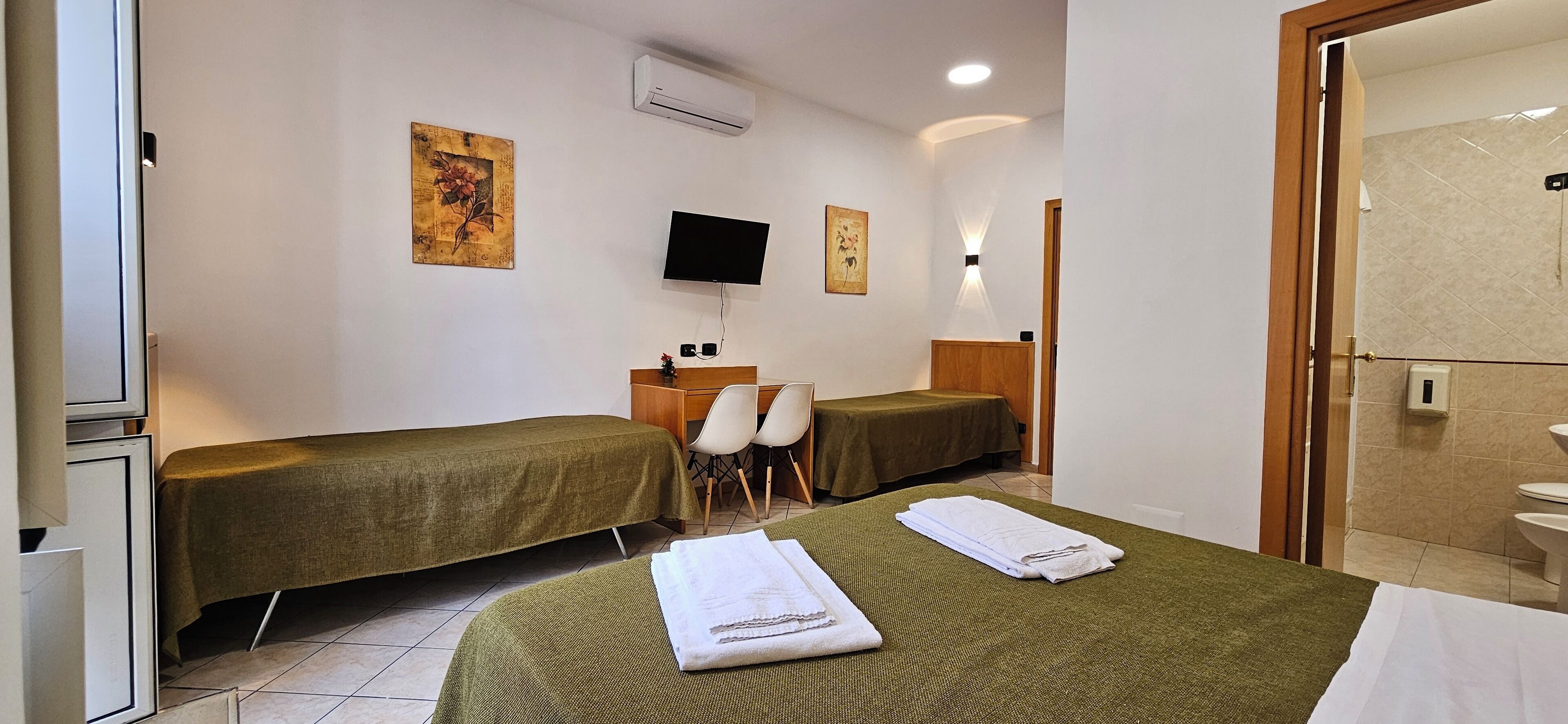 Ein Hotelzimmer mit zwei Einzelbetten, einem Fernseher, einem Schreibtisch mit Stühlen und einem durchgehenden Türrahmen einsehbarer Badezimmer.