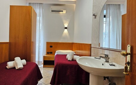 A hotel room with two single beds, a wooden wardrobe, a vanity with a mirror, a sink, and an air conditioning unit.
