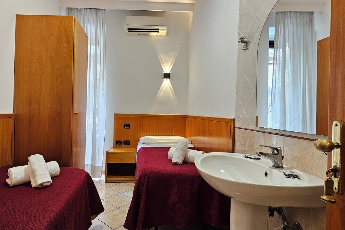A hotel room with two single beds, a wooden wardrobe, a vanity with a sink, and a mirror.