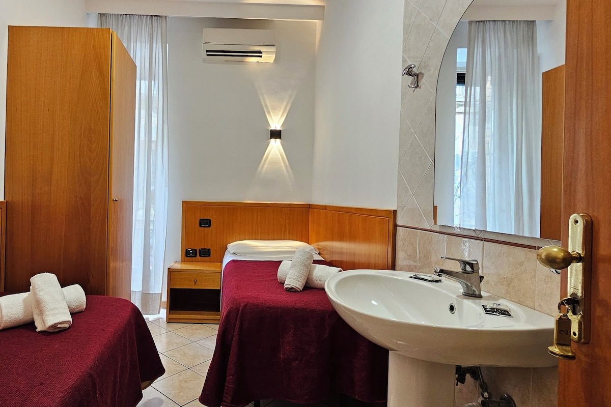 A hotel room with two single beds, a wooden wardrobe, a vanity with a mirror, a sink, and an air conditioning unit.