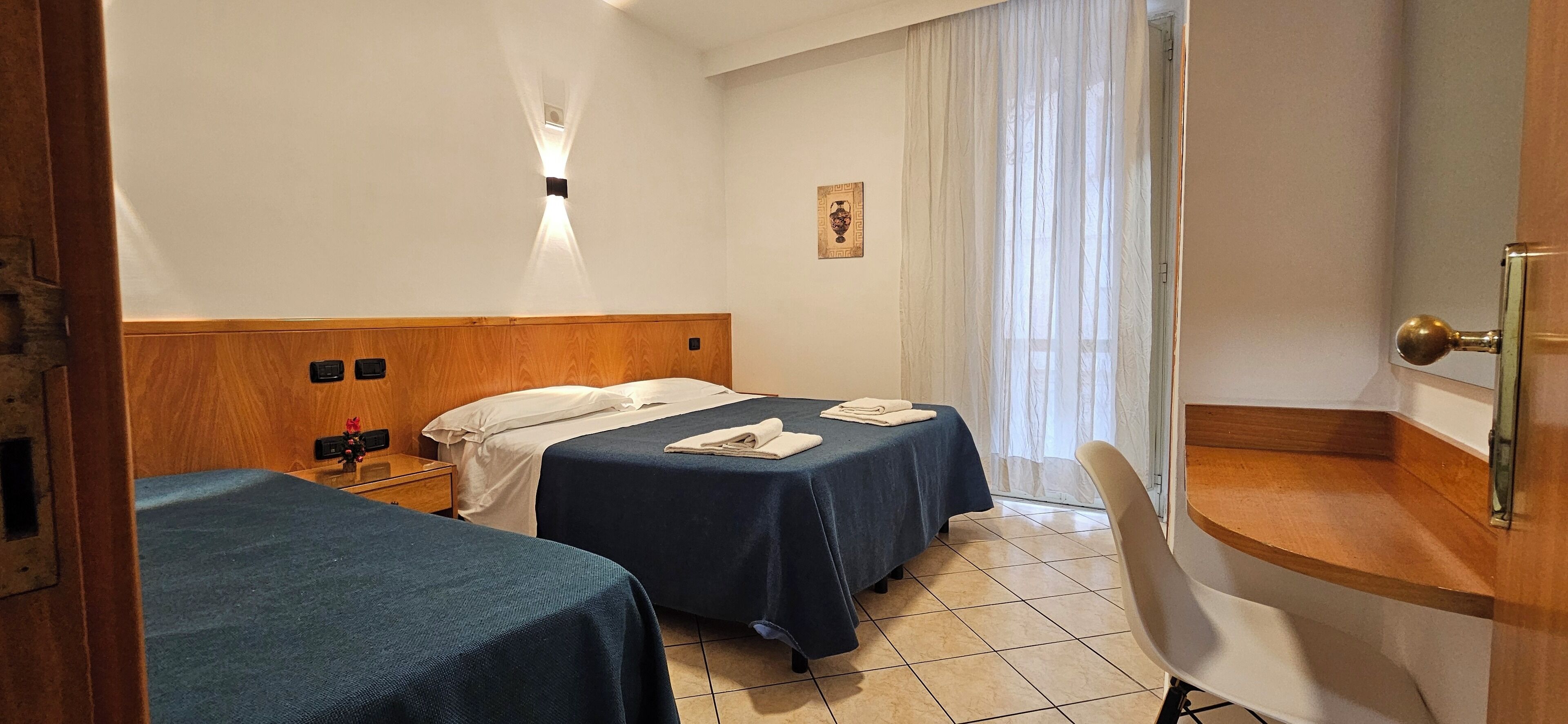Habitación de hotel con dos camas, una mesita de noche, un escritorio, una silla, una ventana con cortinas traslúcidas y un aplique de pared.