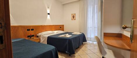 Habitación de hotel con dos camas, una mesita de noche, un escritorio, una silla, una ventana con cortinas traslúcidas y un aplique de pared.