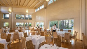 A spacious dining area with multiple tables and chairs, large windows, and a high ceiling with wooden beams. - Ethra Reserve - Alborèa 5* (Castellaneta)