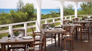 An outdoor dining area with wooden tables and chairs, white railings, and a view of the sea and greenery. - Ethra Reserve - Alborèa 5* (Castellaneta)