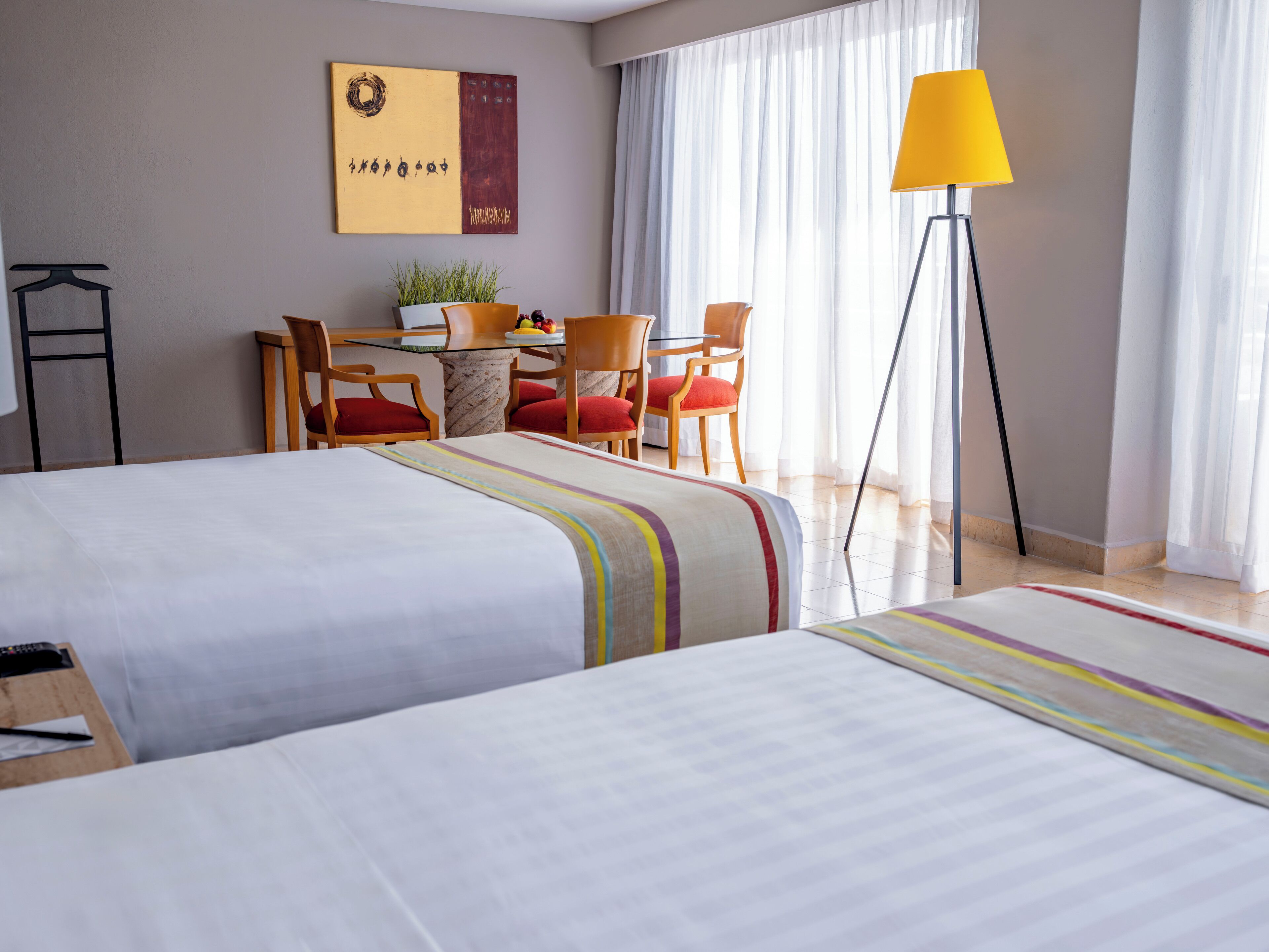 A hotel room with two beds, a desk with chairs, a lamp, and a window with curtains.