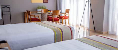 A hotel room with two beds, a desk with chairs, a lamp, and a window with curtains.