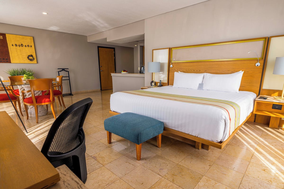 A hotel room with a large bed, a desk with a chair, a small table, and a bench.