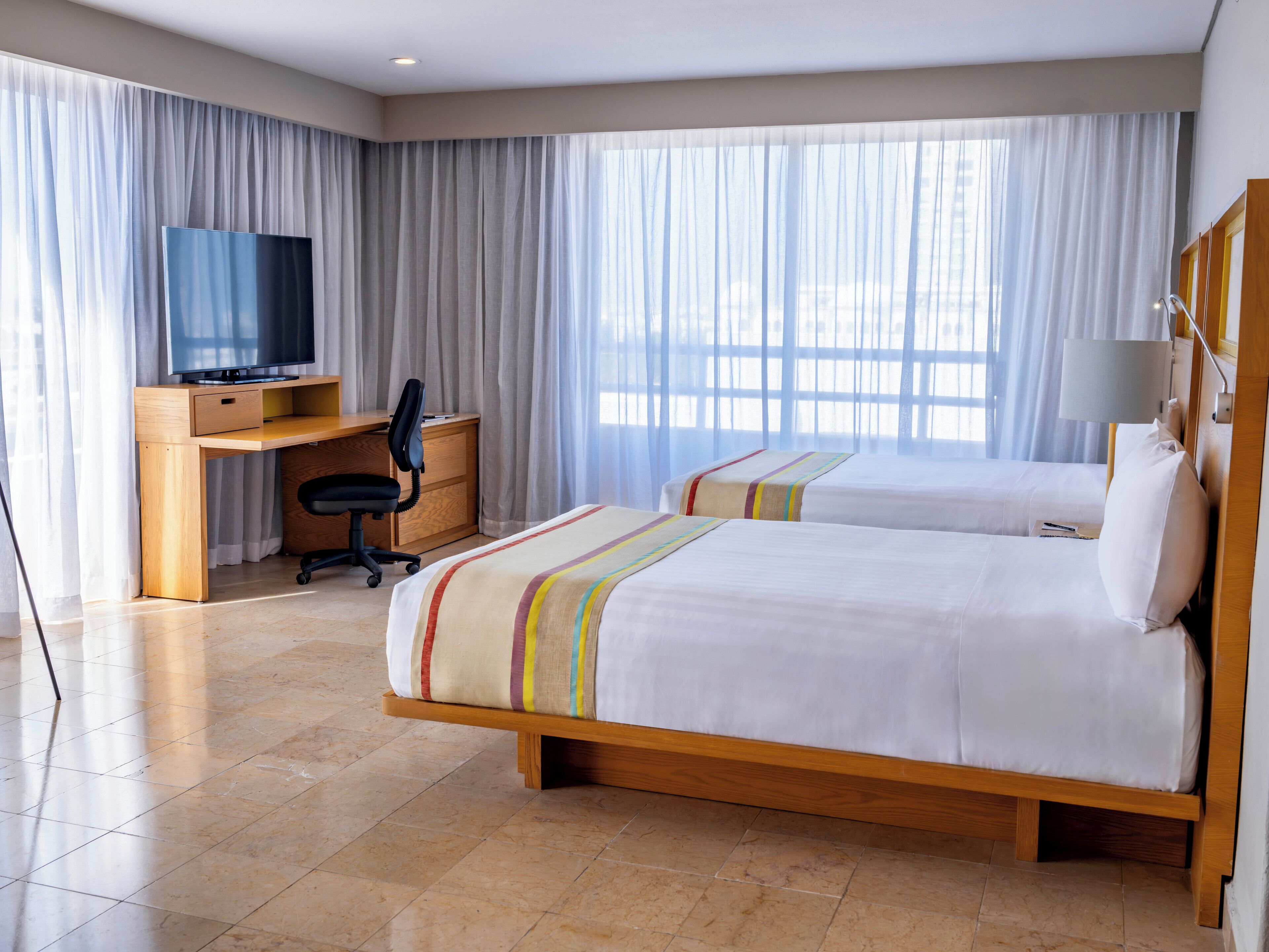 Habitación de hotel con dos camas, un escritorio con silla, televisor y un ventanal con cortinas.