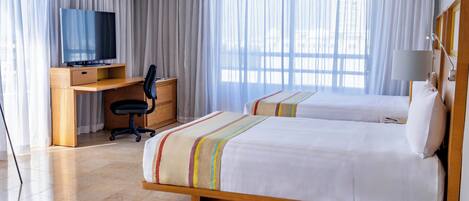 Habitación de hotel con dos camas, un escritorio con silla, televisor y un ventanal con cortinas.