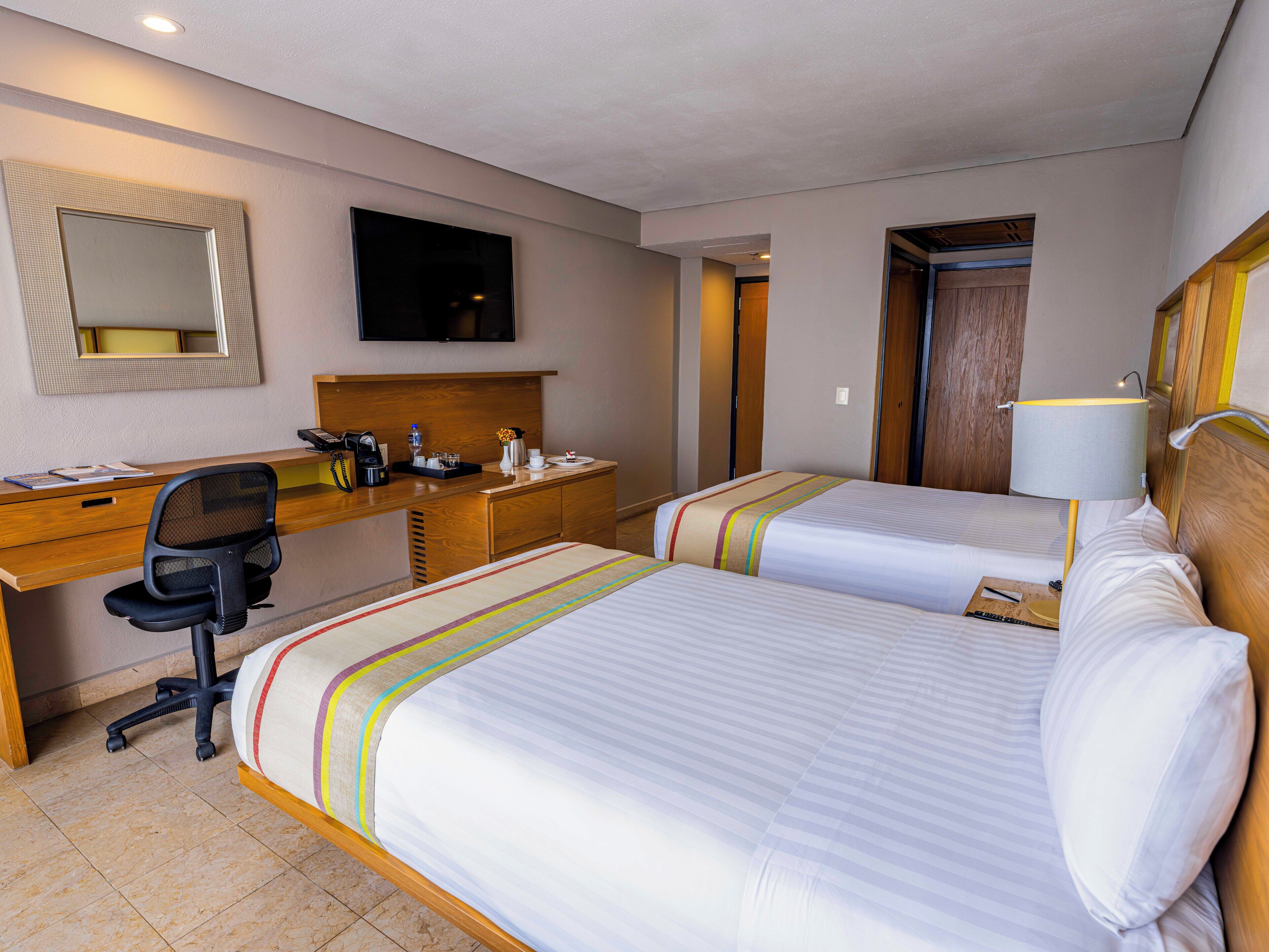 A hotel room with two beds, a desk, a chair, a TV, and a lamp.