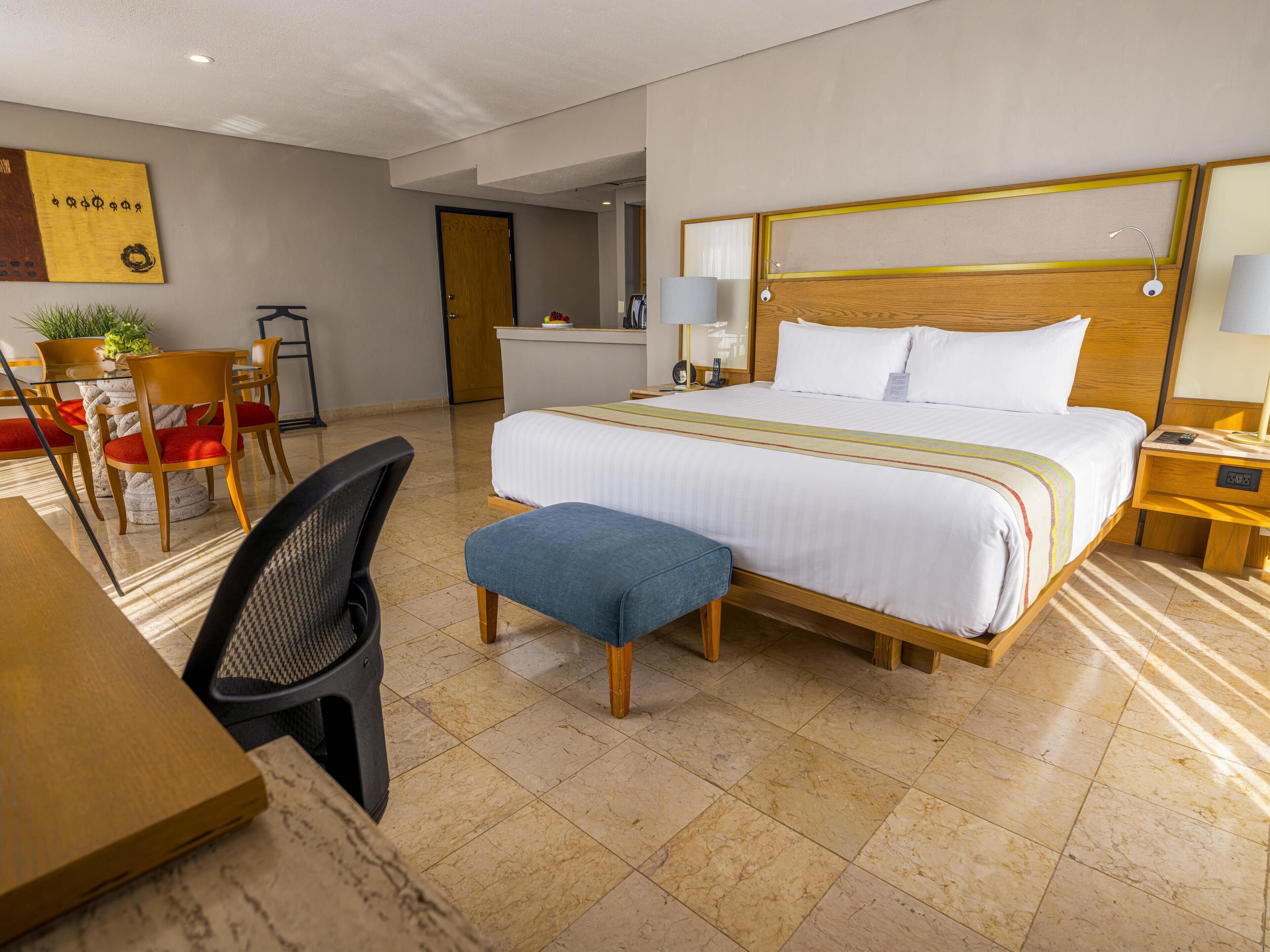 A hotel room with a large bed, a desk with a chair, a small table, and a potted plant.