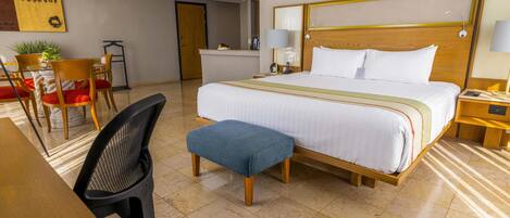 A hotel room with a large bed, a desk with a chair, a small table, and a potted plant.