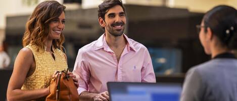 Un mostrador de recepción con una computadora portátil, una mujer con un vestido amarillo y un hombre con una camisa rosa.
