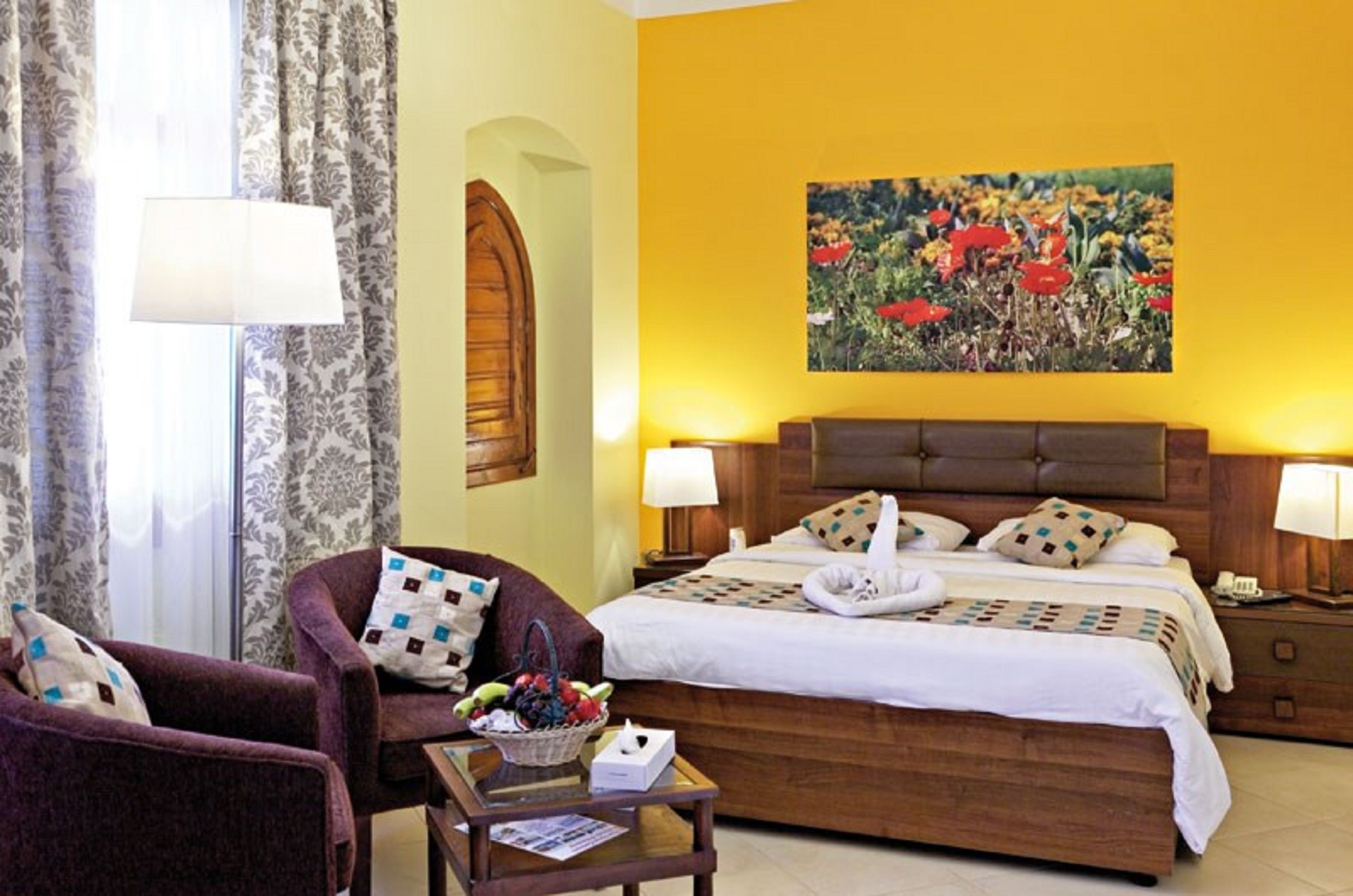 Une chambre d’hôtel avec un grand lit, deux tables de chevet, une chaise, une petite table sur laquelle se trouve un panier de fruits, et un tableau accroché au mur.