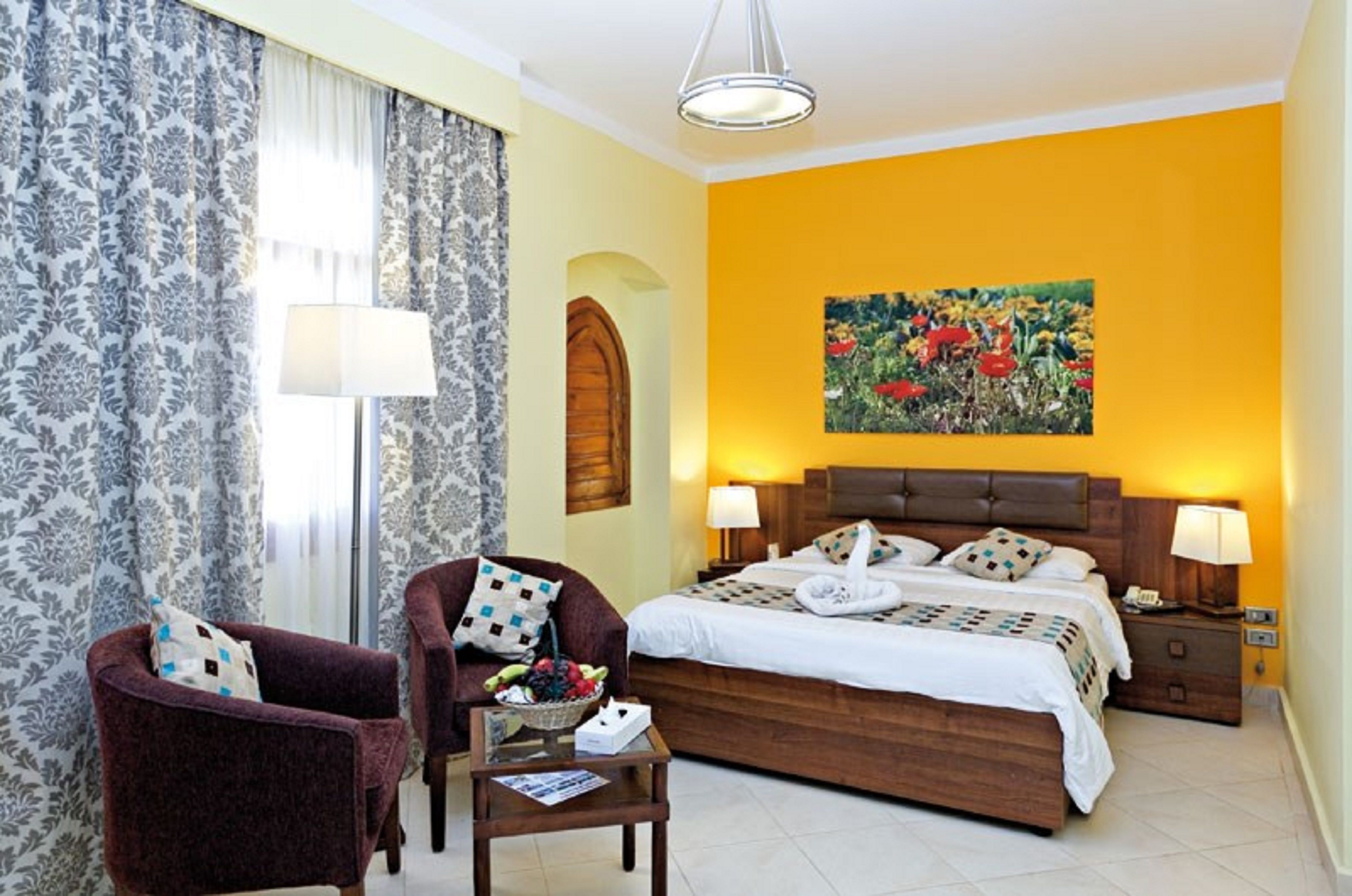 Une chambre d’hôtel avec un lit, deux chaises, une table sur laquelle se trouve un saladier de fruits, un tableau au mur et une lampe.