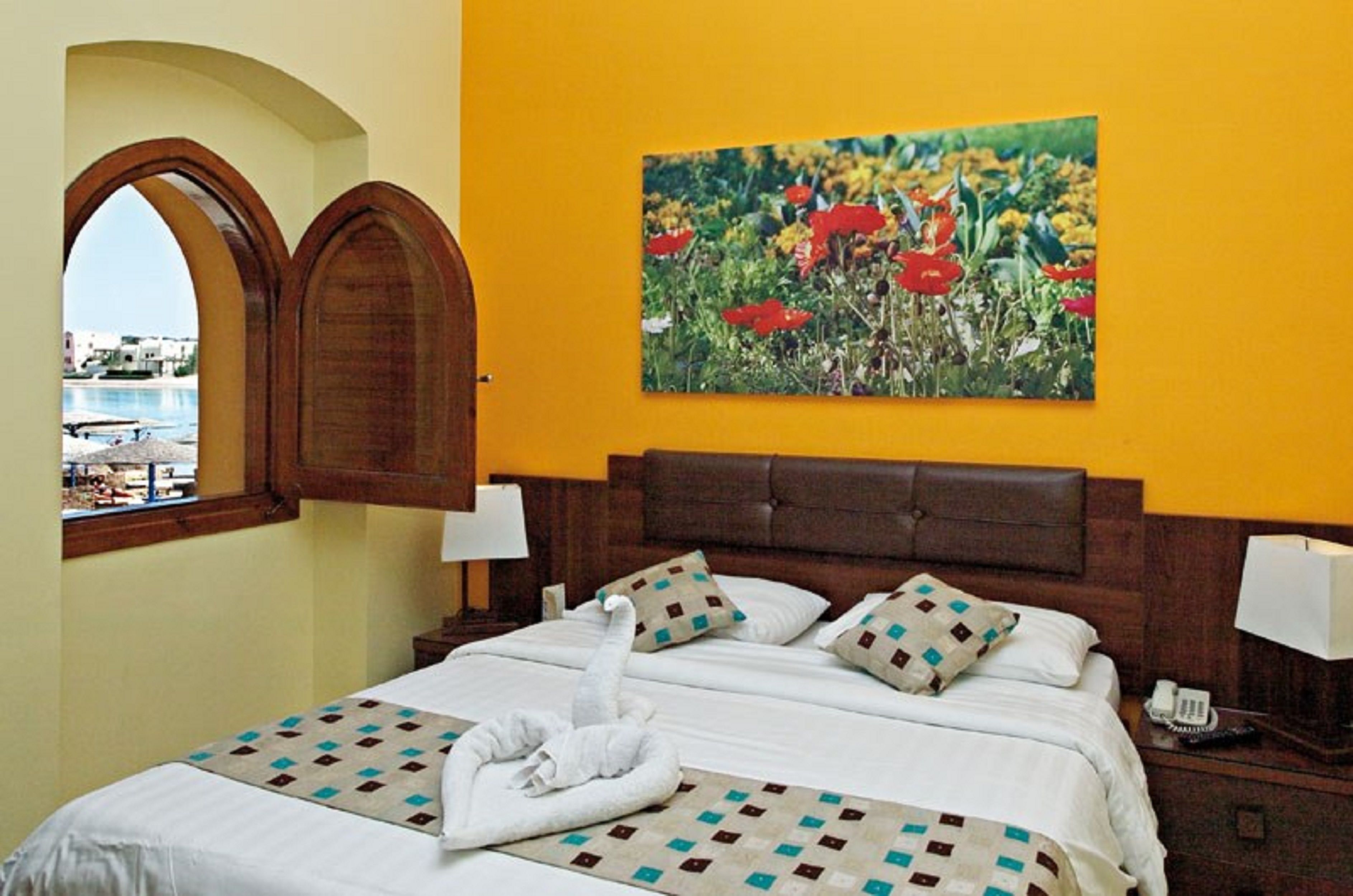 Habitación de hotel con una cama, un cuadro de flores, un ventanal de arco y vistas al mar.