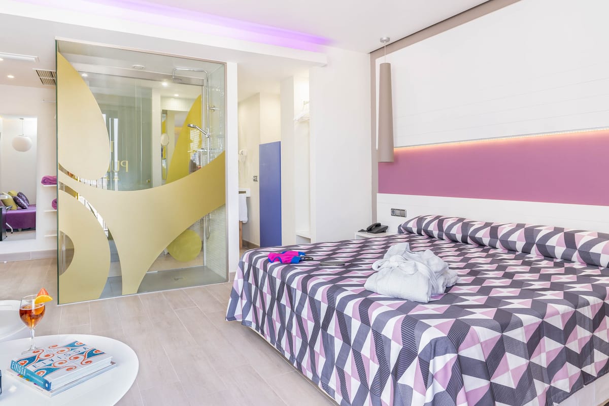 Une chambre d’hôtel moderne avec un grand lit, une douche en verre et une table ronde sur laquelle se trouvent un livre et un verre.