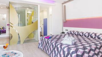 A modern hotel room with a large bed, a glass shower area, and a round table with a book and a glass of drink.