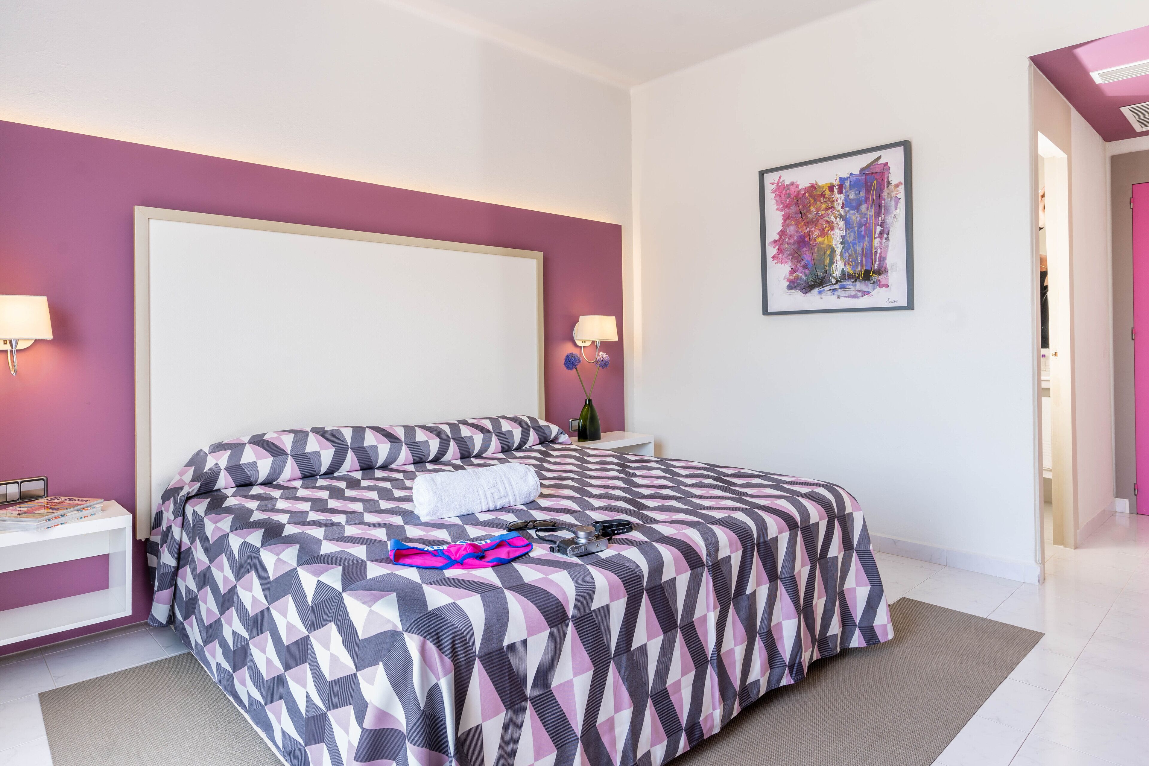 Een slaapkamer met een groot bed, een nachtkastje, een wandlamp en een kunstwerk in een lijst.