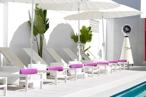 Un área junto a la piscina con sillas reclinables blancas, toallas moradas y sombrillas.