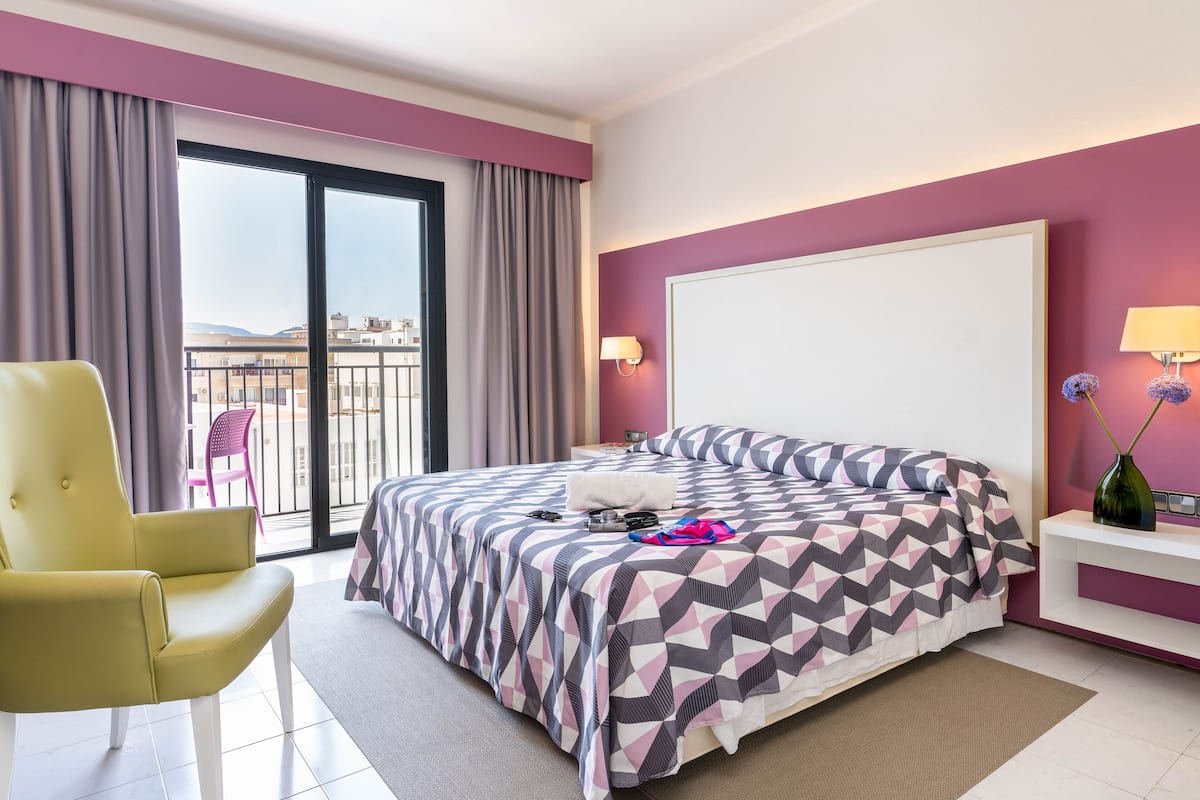Une chambre d’hôtel avec un lit, un fauteuil jaune, une table de chevet et une vue sur la ville par une grande fenêtre.