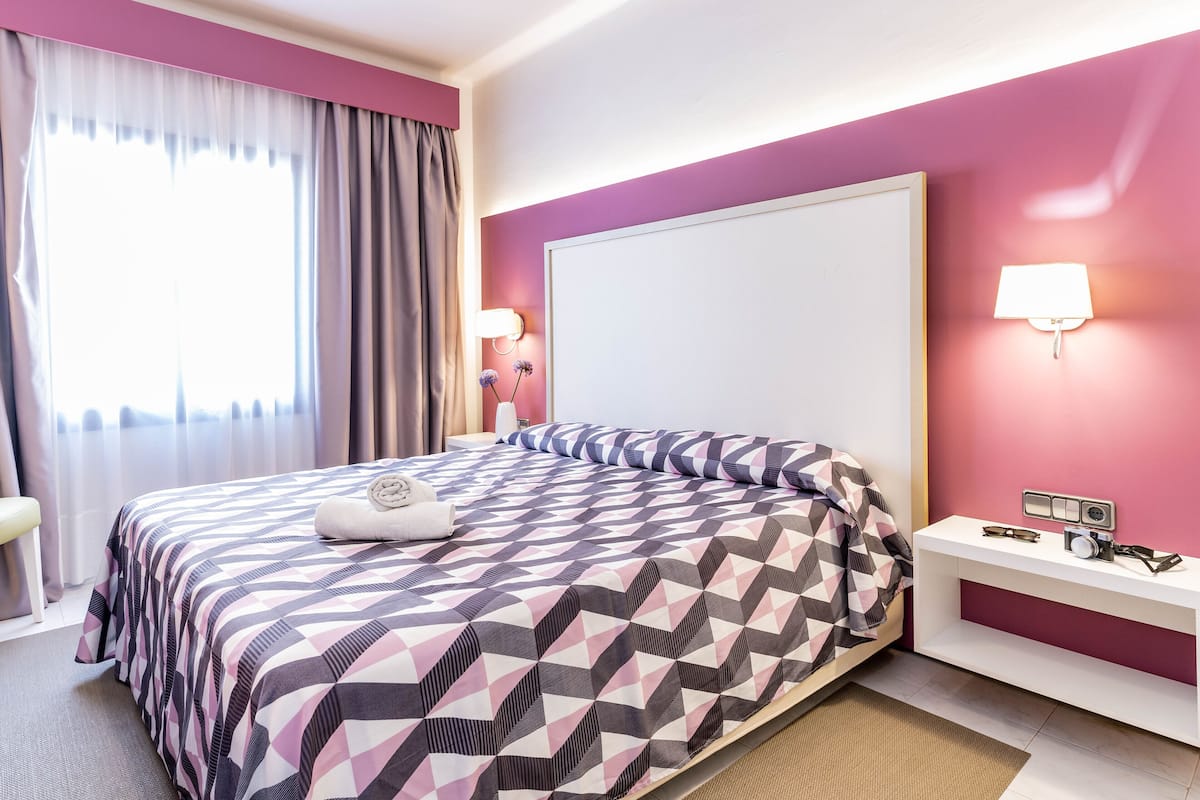 Une chambre d’hôtel moderne avec un grand lit, une table de chevet avec une lampe et une vue sur la ville par la fenêtre.