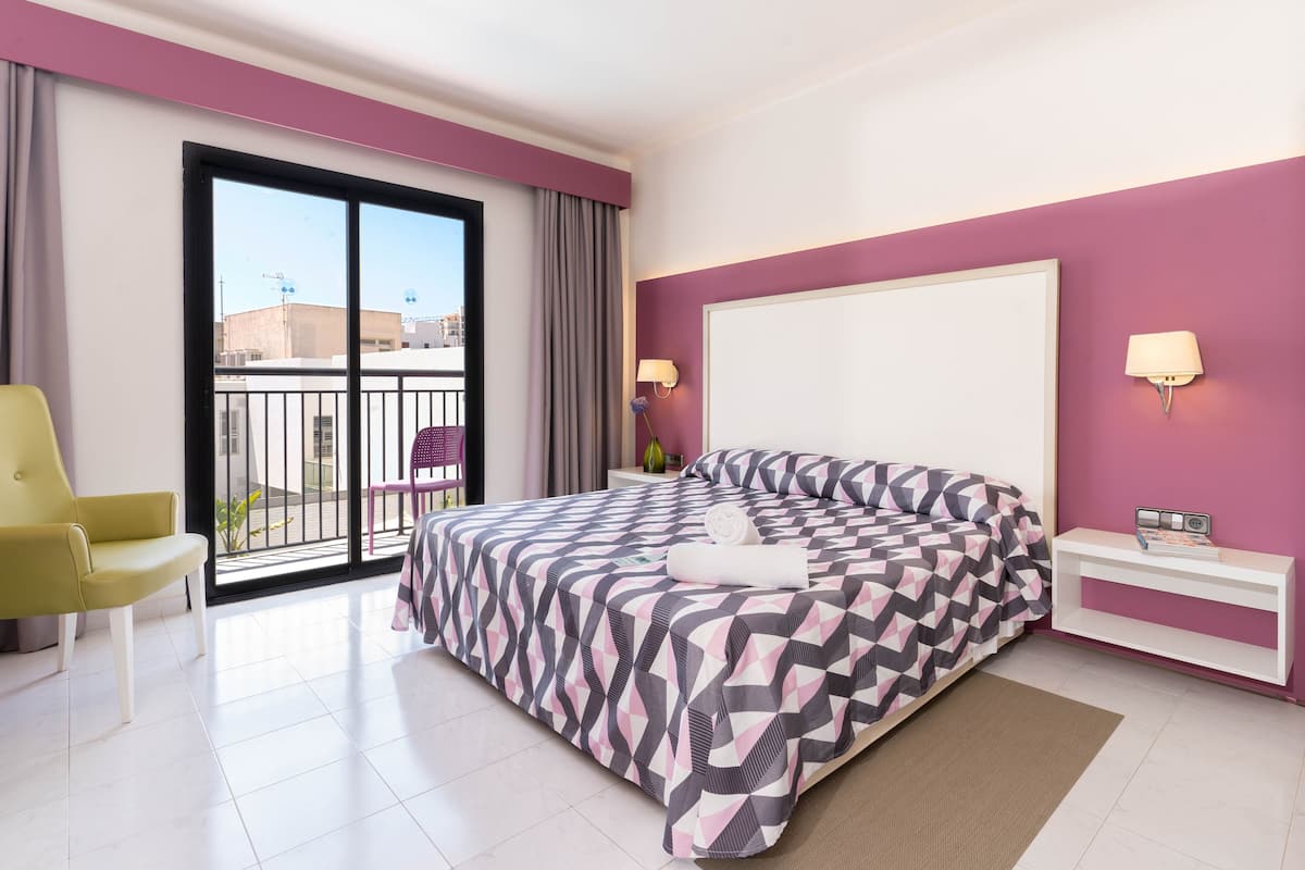 Une chambre d’hôtel moderne avec un grand lit, une chaise jaune, une petite table et un balcon offrant une vue sur les bâtiments.