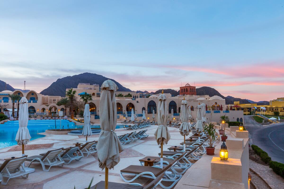 A resort pool area with lounge chairs and umbrellas, a swimming pool, and a view of mountains.