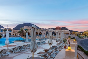 Área de piscina en un resort con sillones de descanso y sombrillas, una alberca y vistas a montañas.