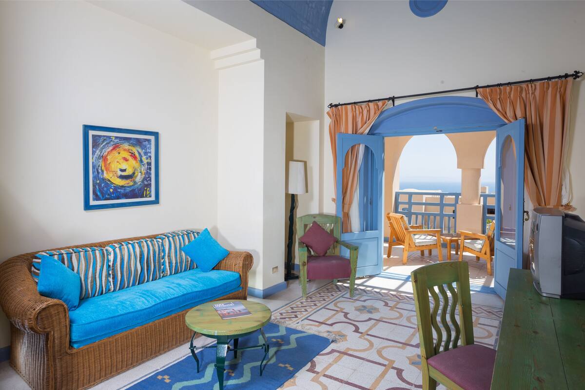A cozy living room with a wicker sofa, a small round table, and a chair. There is a painting on the wall and a view of the sea through an open doorway.